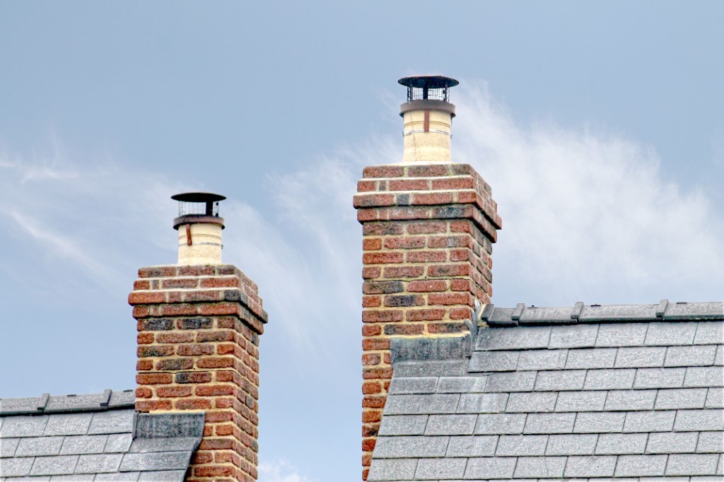 Two brick chimneys with cream pots and metal cowls on a slate roof under a bright sky